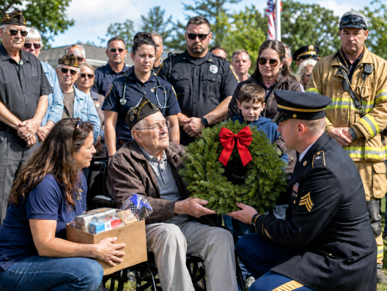 Elderly veteran in a wheelchair receiving a green remembrance wreath with a red bow from a uniformed servicemember, surrounded by first responders, veterans, and community members outdoors.